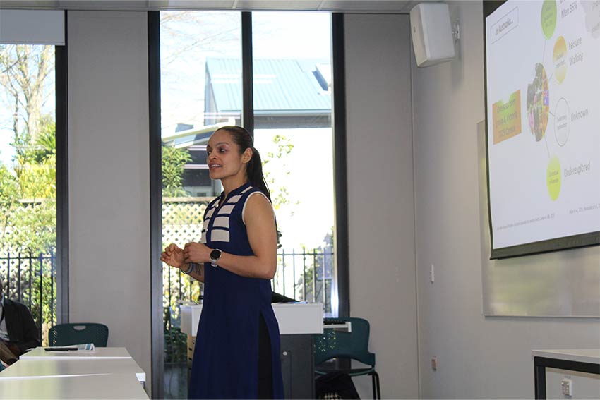 A speaker presents in a seminar room, standing beside a projected slide showing a diagram with labelled circles.